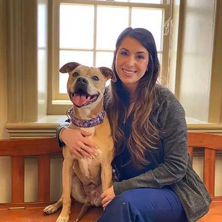 A woman smiling with her tan dog, both seated by a window.