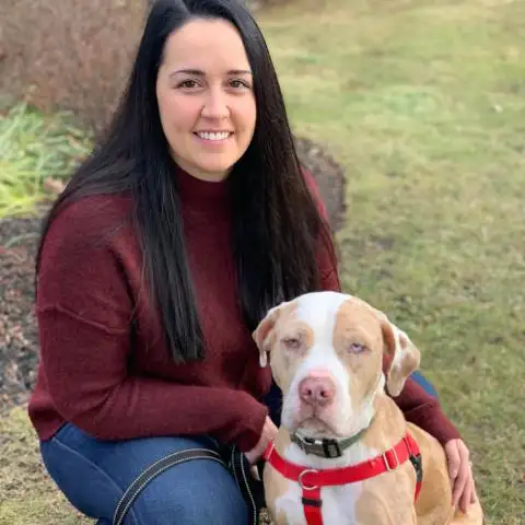 A woman in a maroon sweater smiling beside a seated brown and white dog with a red collar.